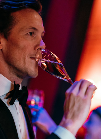 a man in a tuxedo drinking a glass of wine.